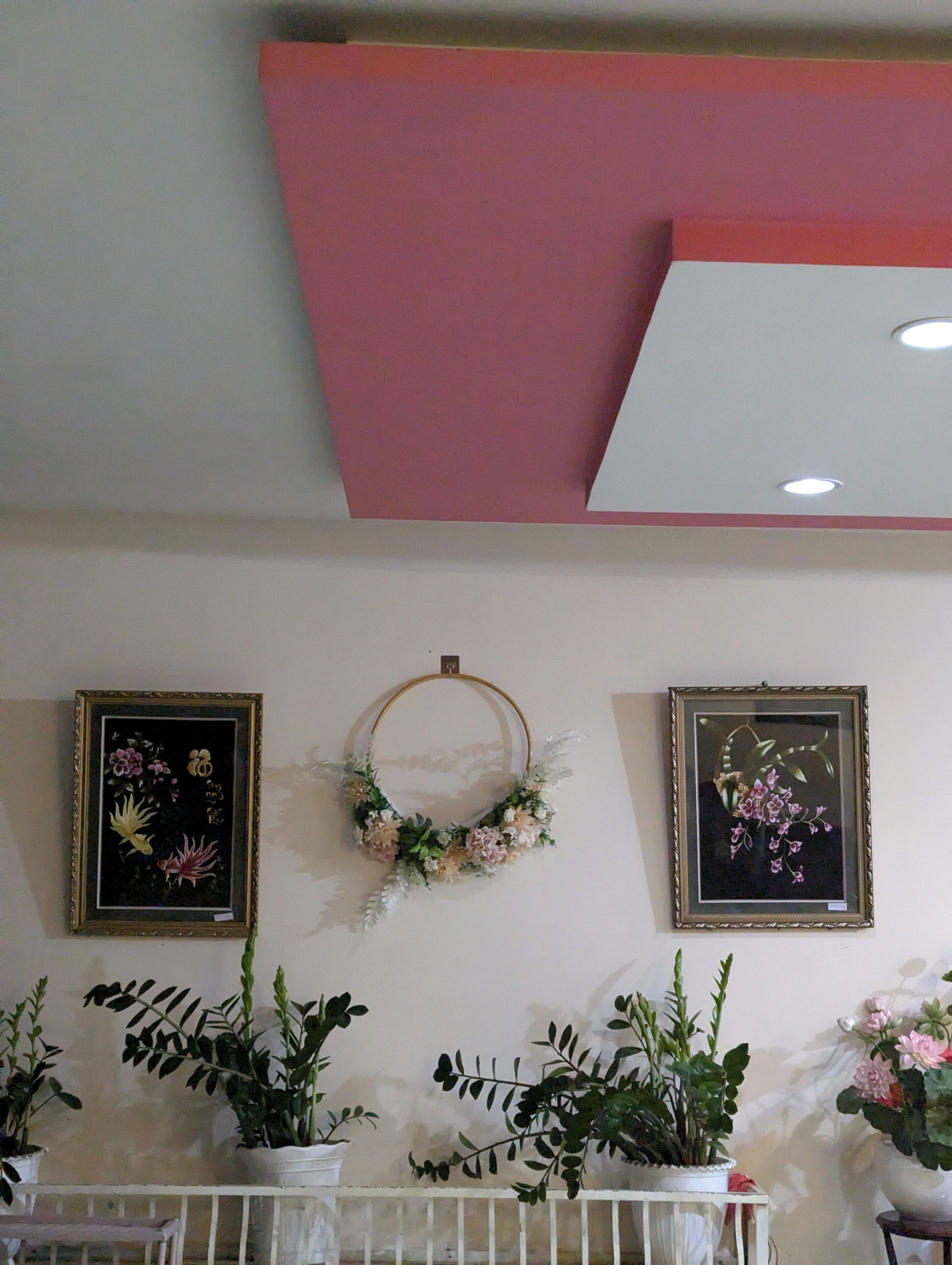 A room with a white ceiling, a pink accent wall, and a white railing. There are two framed floral artworks on the wall, a wreath hanging on the ceiling, and several potted plants on the railing.