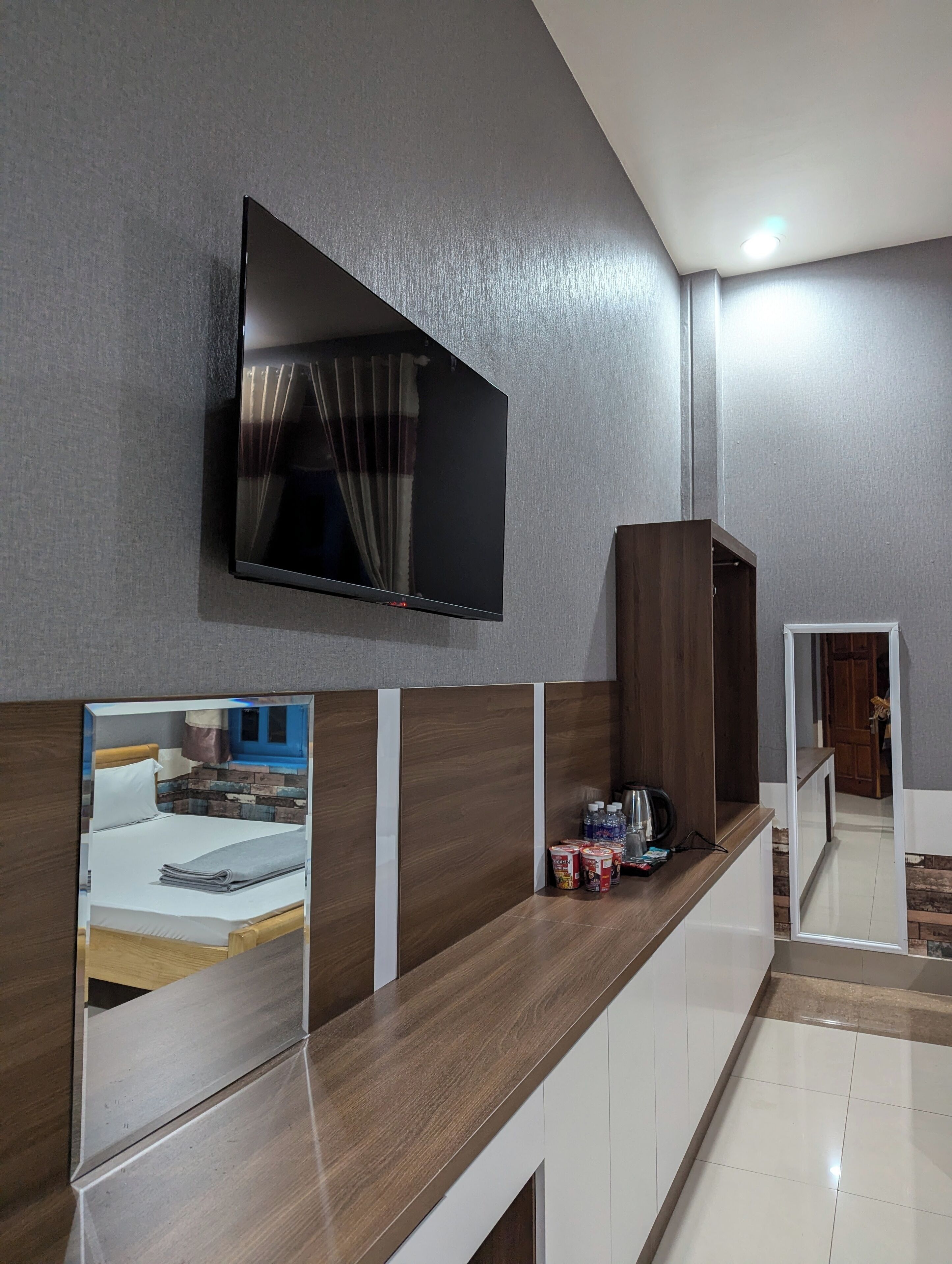 A modern hotel room with a large flat-screen TV mounted on the wall, a wooden bench with a mirror, and a small table with toiletries.