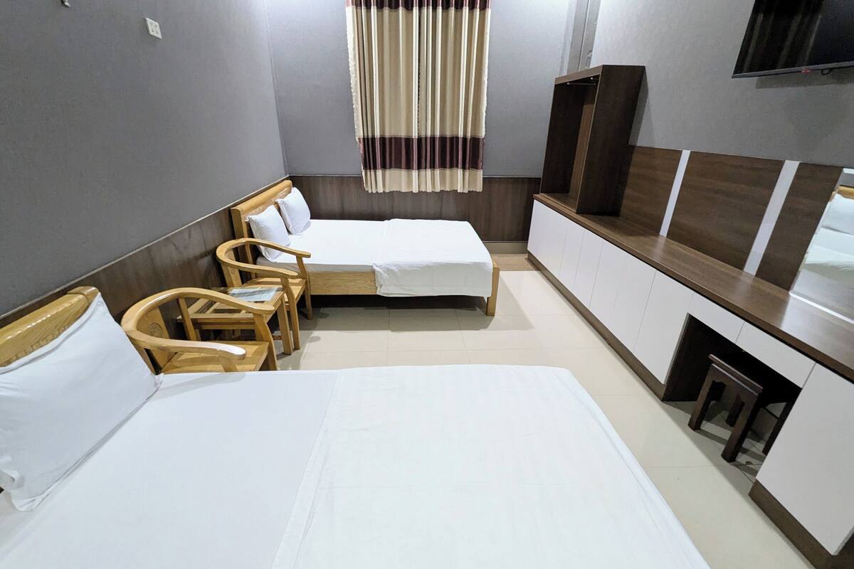 Quarto de hotel com duas camas de solteiro, uma cadeira de madeira, uma mesa pequena, uma televisão e um armário.