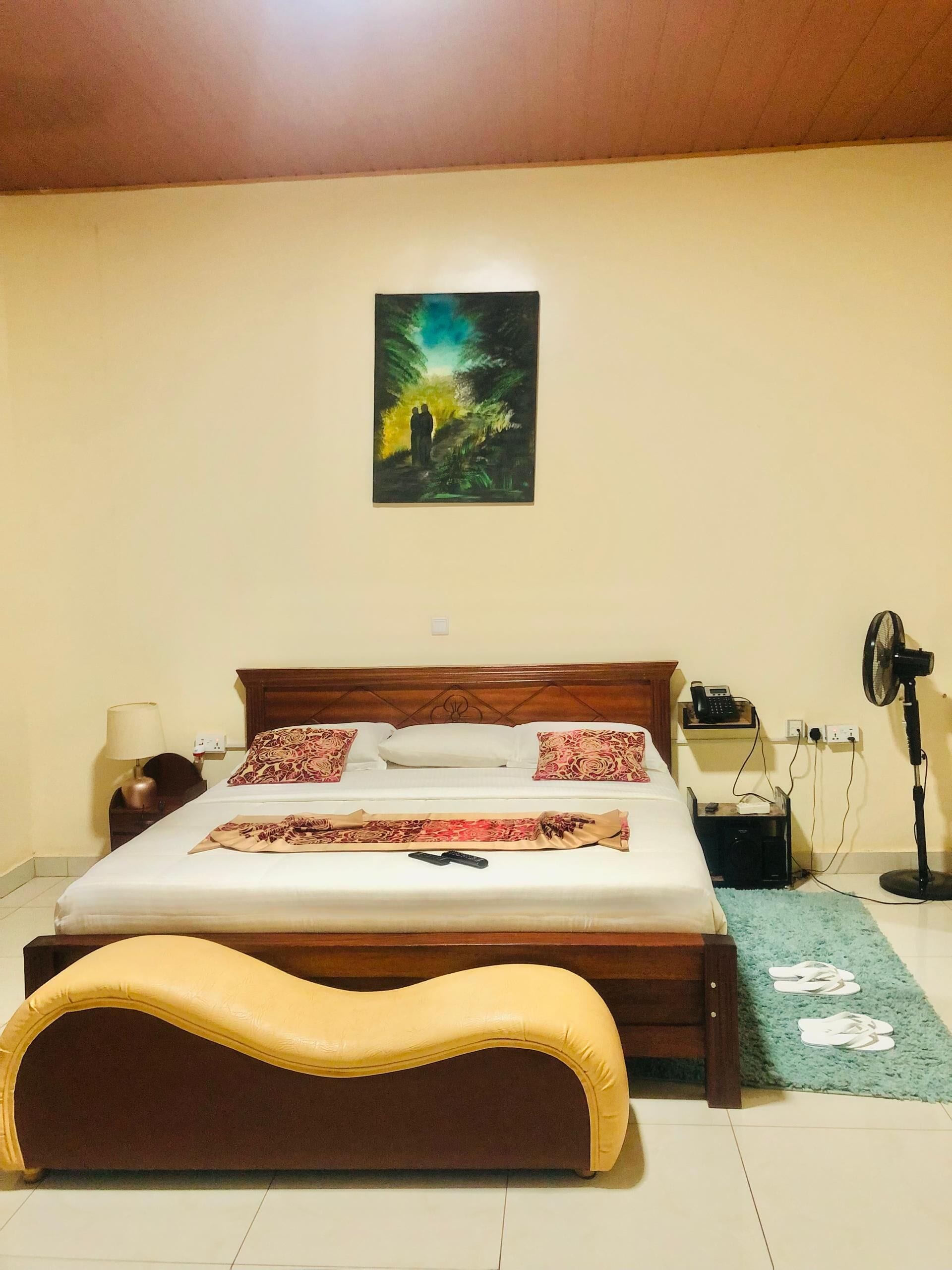 A bedroom with a bed, bedside table, lamp, phone, fan, and slippers on a rug.