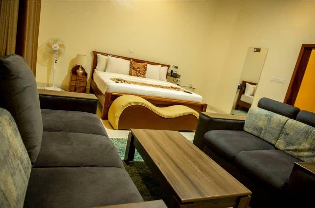 A hotel room with a bed, two sofas, a wooden coffee table, a floor lamp, and a fan.