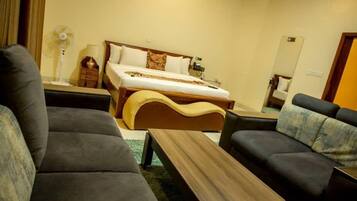 A hotel room with a bed, two sofas, a wooden coffee table, a floor lamp, and a fan.