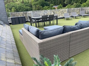 En tagterrasse med en fletsofa, et spisebord med stole og udsigt til et skovområde.