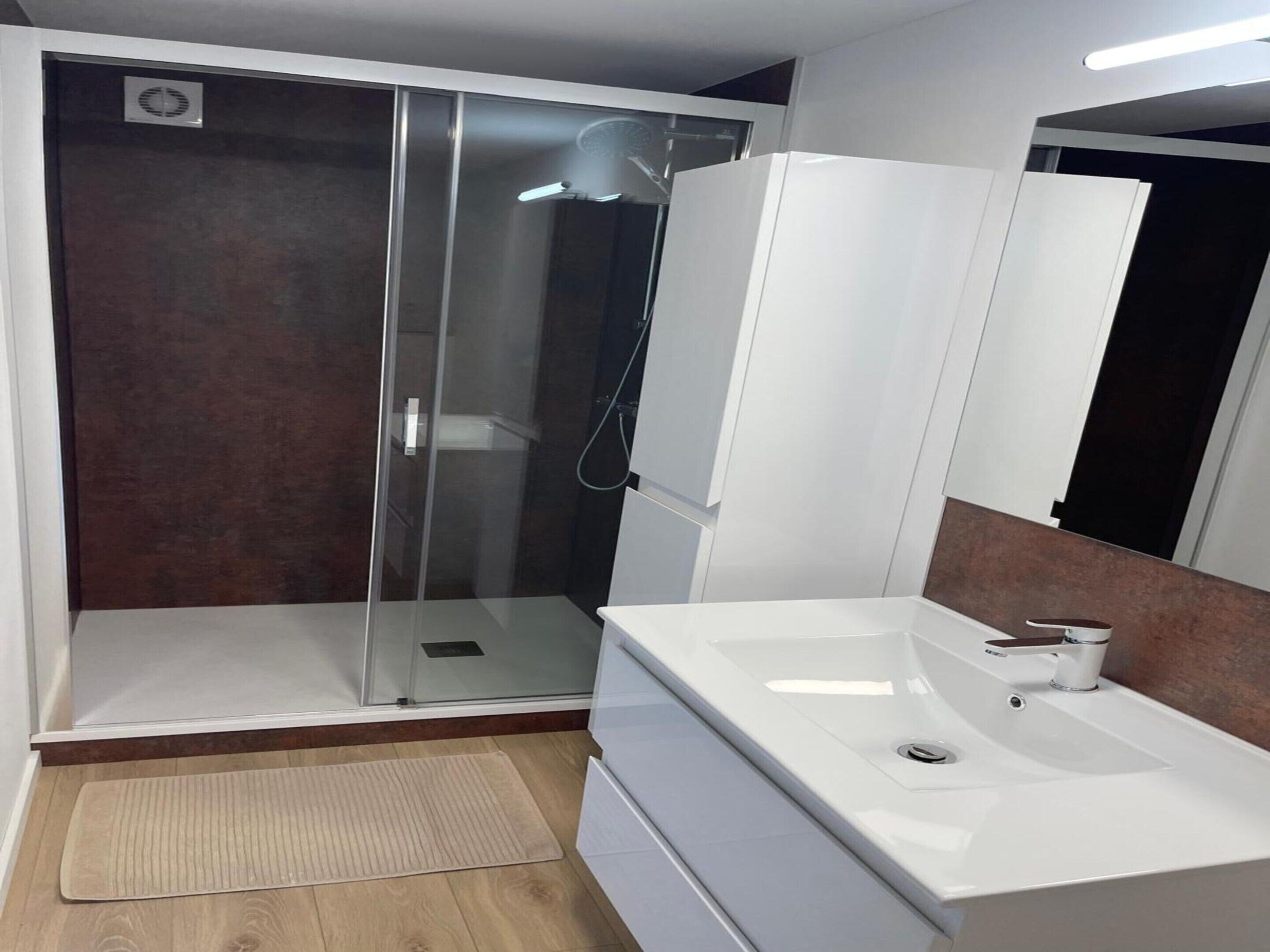 A modern bathroom with a glass shower enclosure, a white sink with a mirror, and a wooden floor.