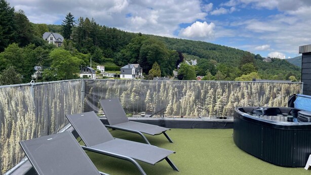 Eine Dachterrasse mit Liegestühlen und Whirlpool, umgeben von einem Metallzaun und Blick auf einen bewaldeten Hang.