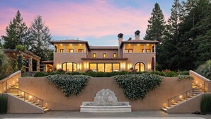 A two-story house with a swimming pool in front, surrounded by lush greenery and a stone fountain.