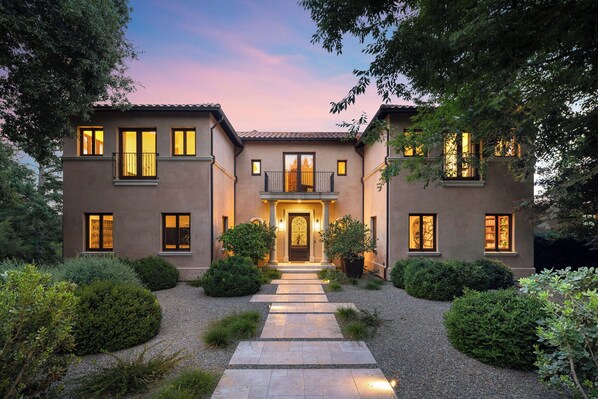 A two-story house with a central entrance, surrounded by well-maintained gardens and a gravel pathway leading to the door.