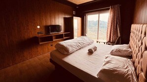 Een hotelkamer met een houten plafond, een bed met witte lakens, een televisie op een houten plank en een raam met gordijnen.