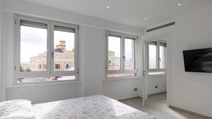 Un dormitorio con cama, ventanas amplias y vistas a edificios.