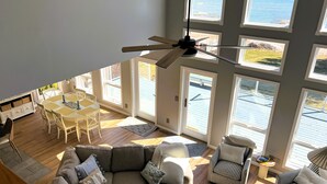 A spacious living room with a high ceiling, large windows, a ceiling fan, a dining area with a table and chairs, and a cozy seating area with a couch and armchairs.