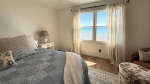 A bedroom with a bed, a nightstand, a lamp, a chair, a window with curtains, and a view of the sea.