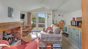 A cozy living room with a fireplace, a sofa, a dining table, and a kitchen area. - The Bothy - North East Escapes (Alnwick)