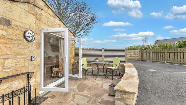 A stone building with a patio area featuring a table and chairs, a door leading to an interior room, and a stone wall. - The Bothy - North East Escapes (Alnwick)
