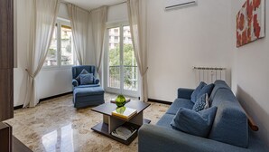 A modern living room with a sofa, armchair, and a coffee table. There is a window with curtains, an air conditioning unit, and a radiator.