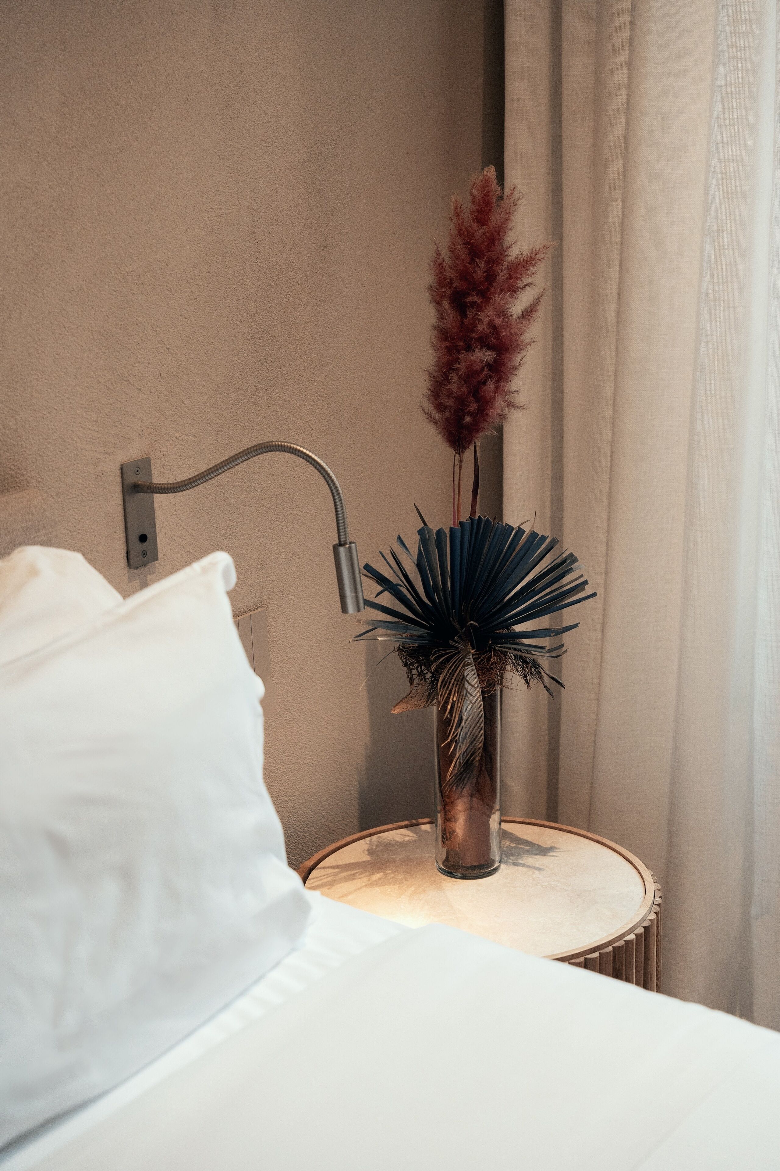 A neatly made bed with white linens, a bedside table with a vase of dried plants, and a wall-mounted lamp.