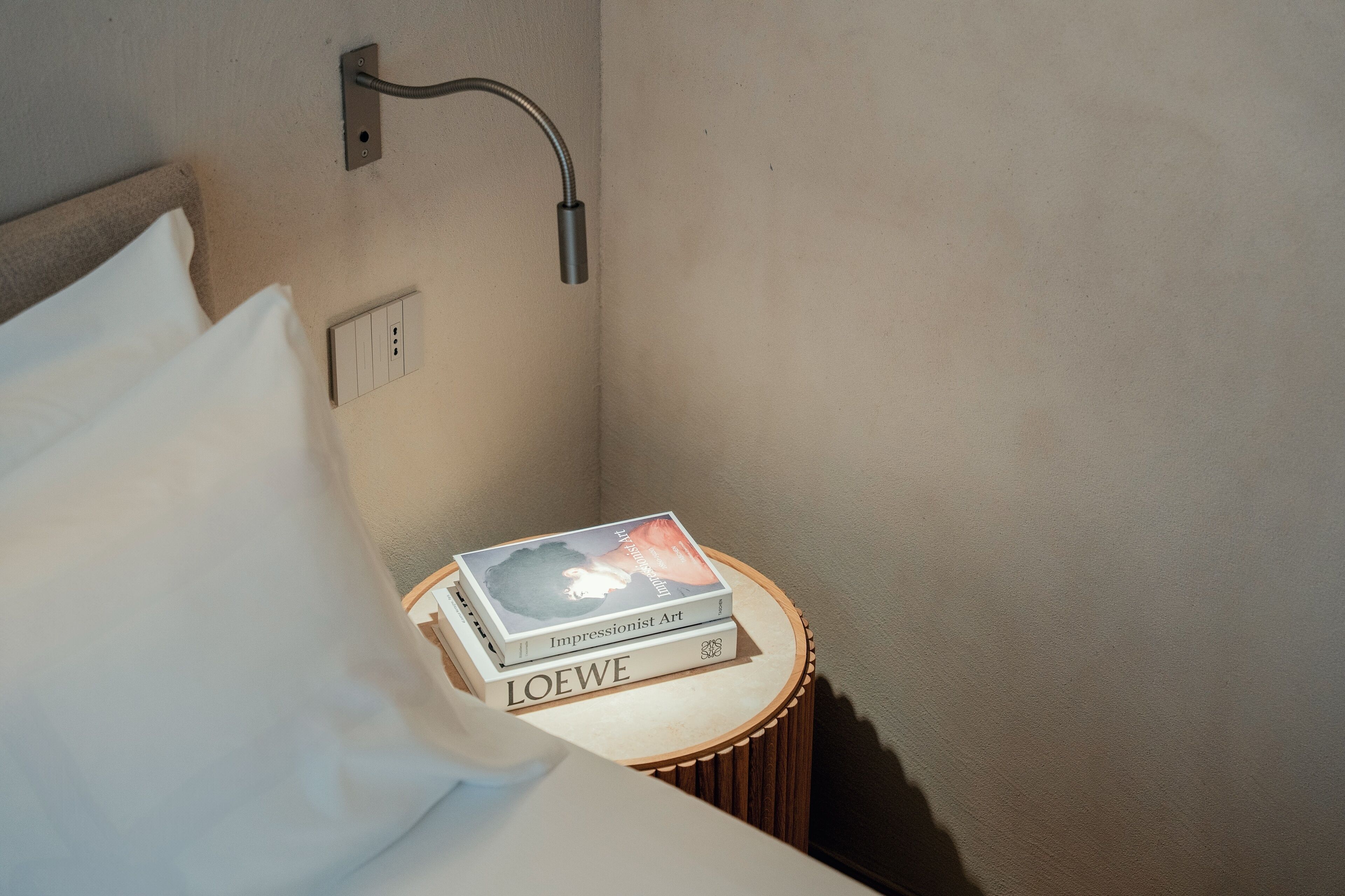 Un lit bien fait, avec un oreiller, une table de chevet sur laquelle reposent deux livres et un luminaire fixé au mur.