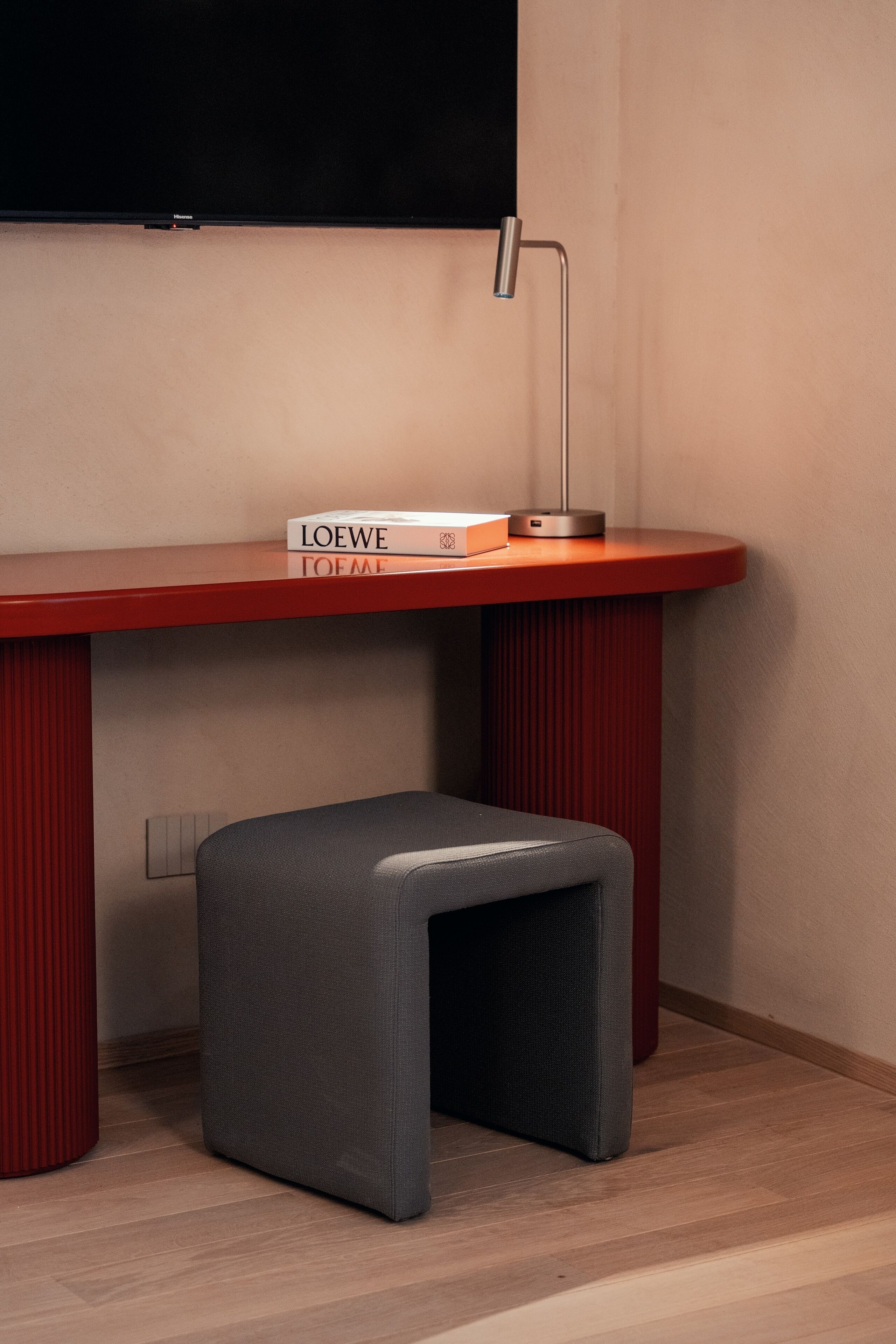 A modern desk with a red finish, a grey ottoman, and a book titled "LOEWE TOFME" on top.