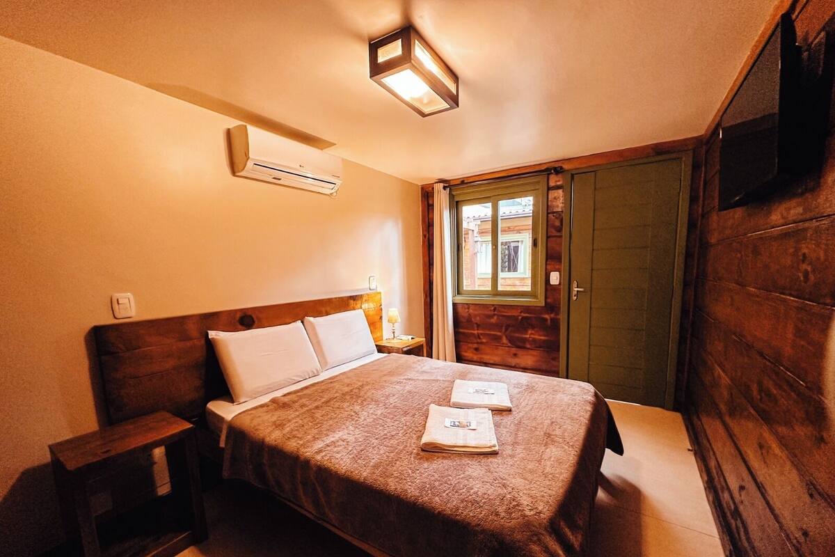 A bedroom with a wooden headboard, a bed with a brown blanket, a bedside table, an air conditioning unit, and a window with a view of the outside.