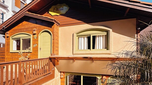 A two-story wooden house with a balcony, a green door, and a small garden area with potted plants.