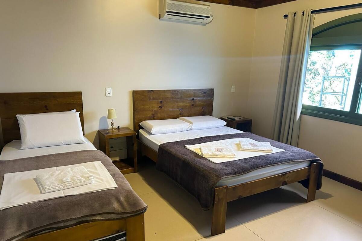 A room with two wooden beds, each with a headboard, a nightstand, and a lamp. There is an air conditioning unit on the wall, and a window with curtains.