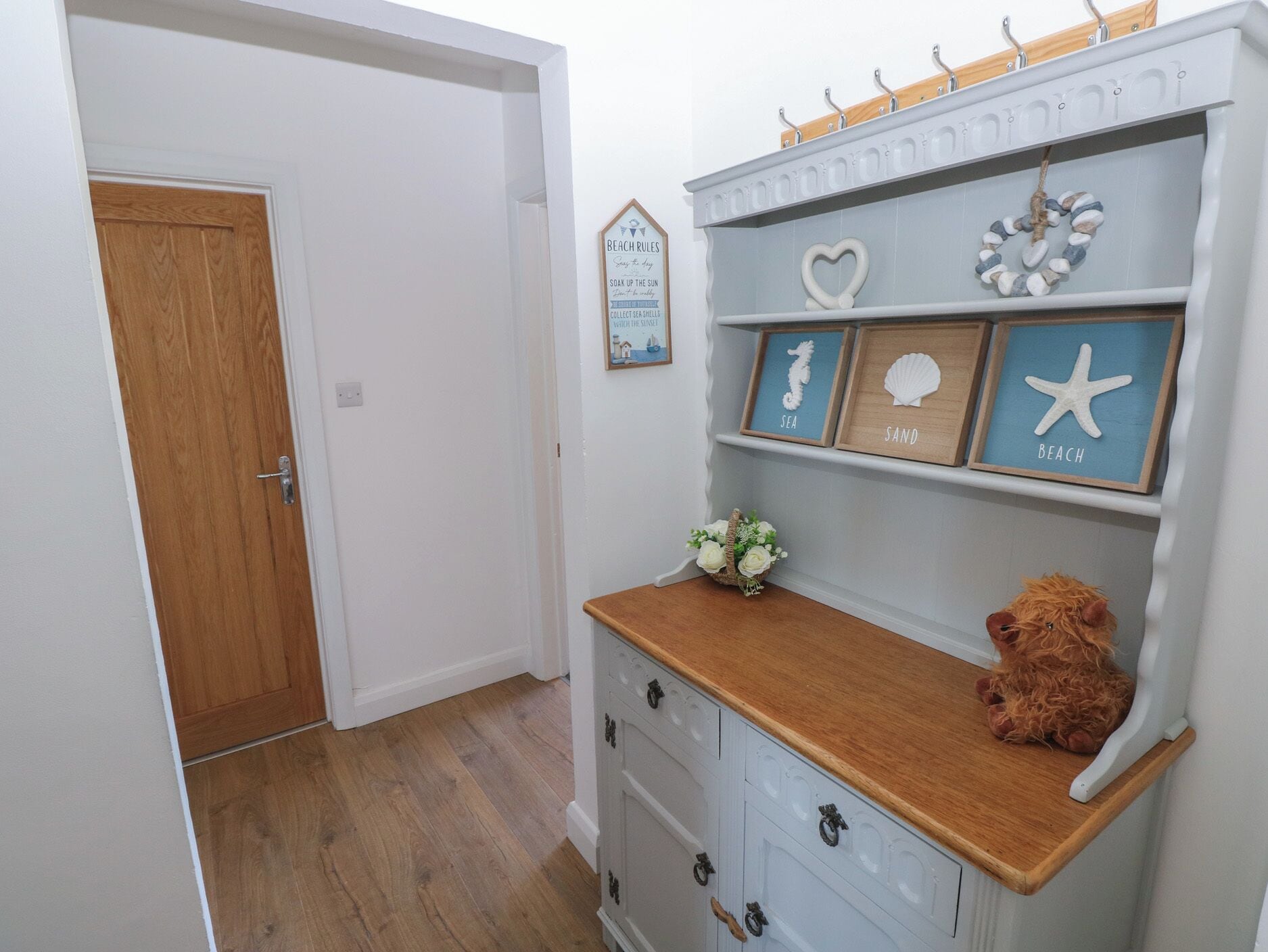 Een gang met een houten deur, een witte muur en een plank met omlijste items, gelabeld 'SEA', 'SAND' en 'BEACH'.