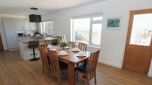 A modern kitchen with white cabinets, a wooden dining table set for six, and a large window providing natural light. - Arfryn (Haverfordwest)