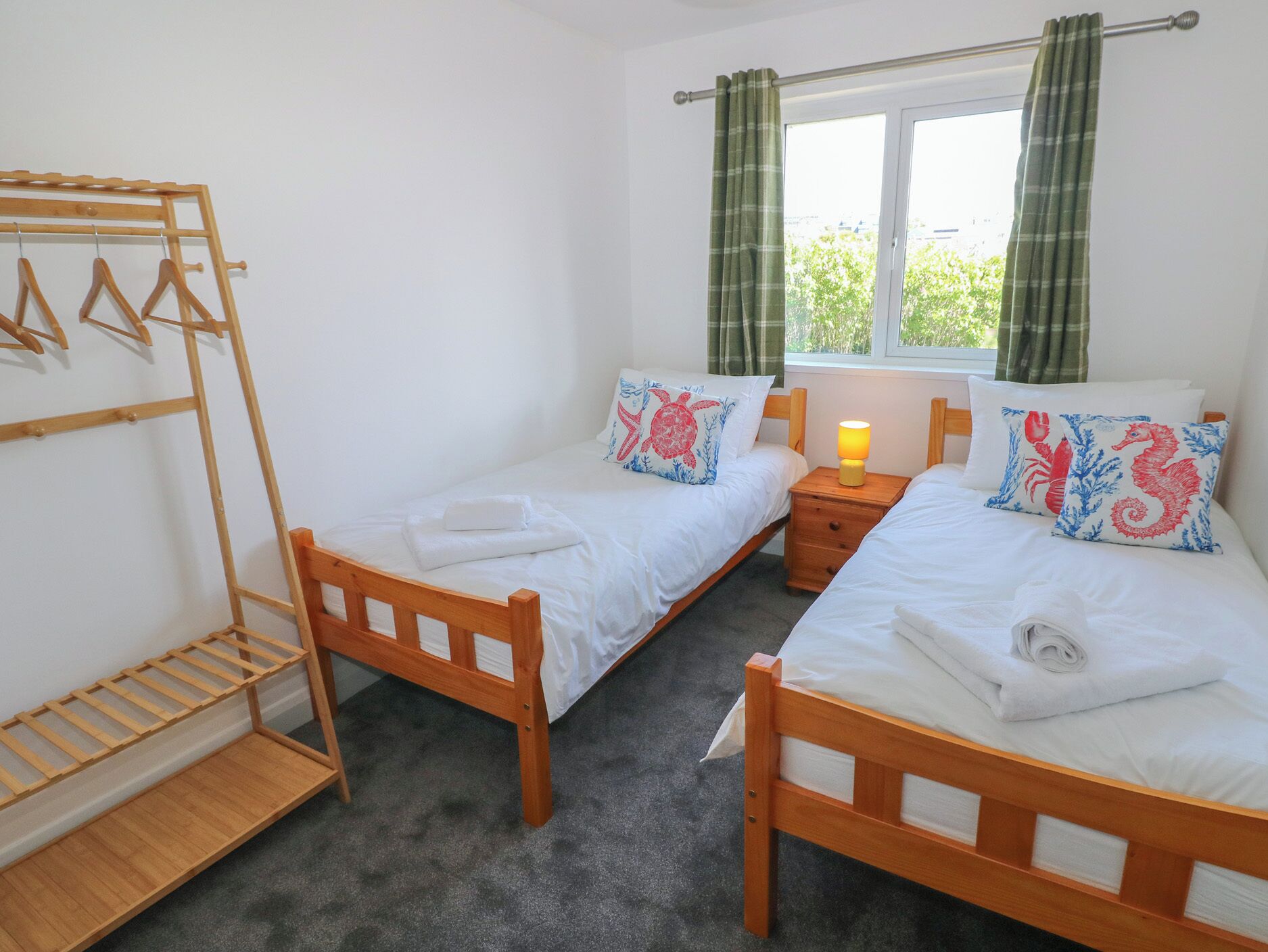 A bedroom with two single beds, a wooden drying rack, a bedside table with a lamp, and a window with curtains.