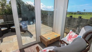 A sunlit patio with a wicker sofa, a wooden coffee table, and a view of a grassy field through large glass doors. - Arfryn (Haverfordwest)
