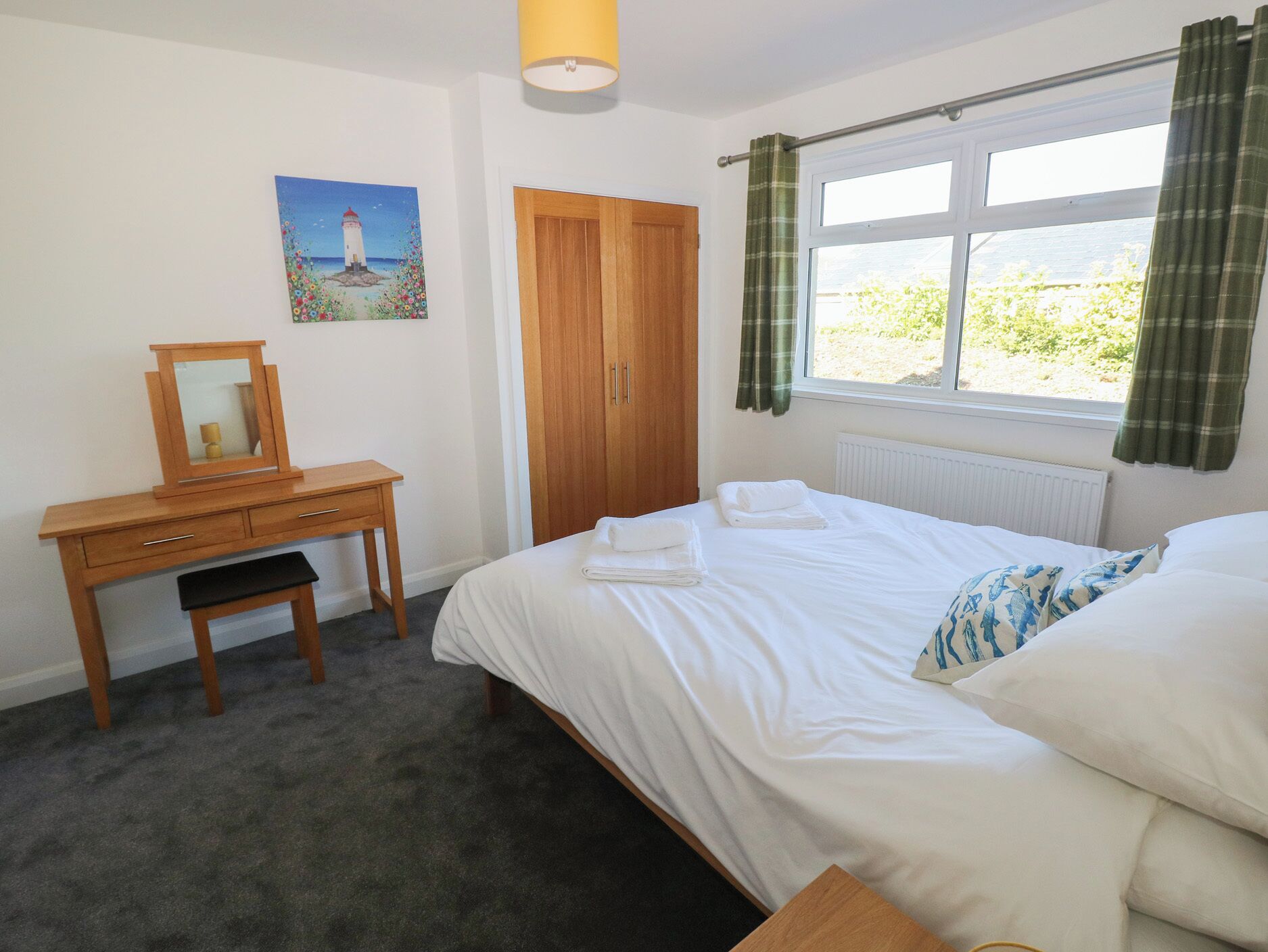 A bedroom with a bed, a wooden desk with a mirror, a painting of a lighthouse, a window with curtains, and a radiator.