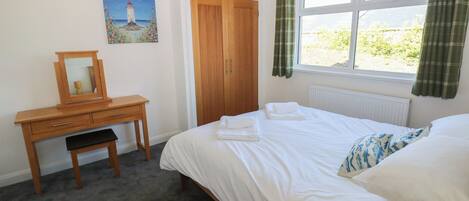 A bedroom with a bed, a wooden desk with a mirror, a painting of a lighthouse, a window with curtains, and a radiator.