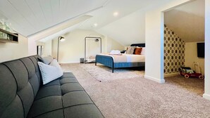 Un dormitorio con techo estilo loft, una cama con colcha azul, un sofá gris oscuro con cojines y una alfombra estampada.