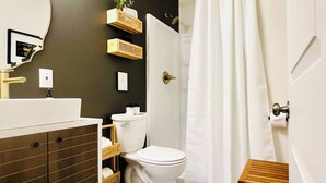 Un baño con inodoro blanco, tocador con espejo, cortina de ducha y un taburete de madera pequeño.