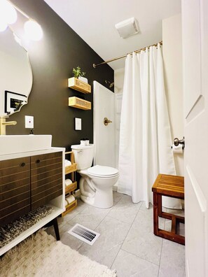 Un bagno con il water bianco, un lavabo con specchio, una tenda da doccia e uno sgabello di legno.
