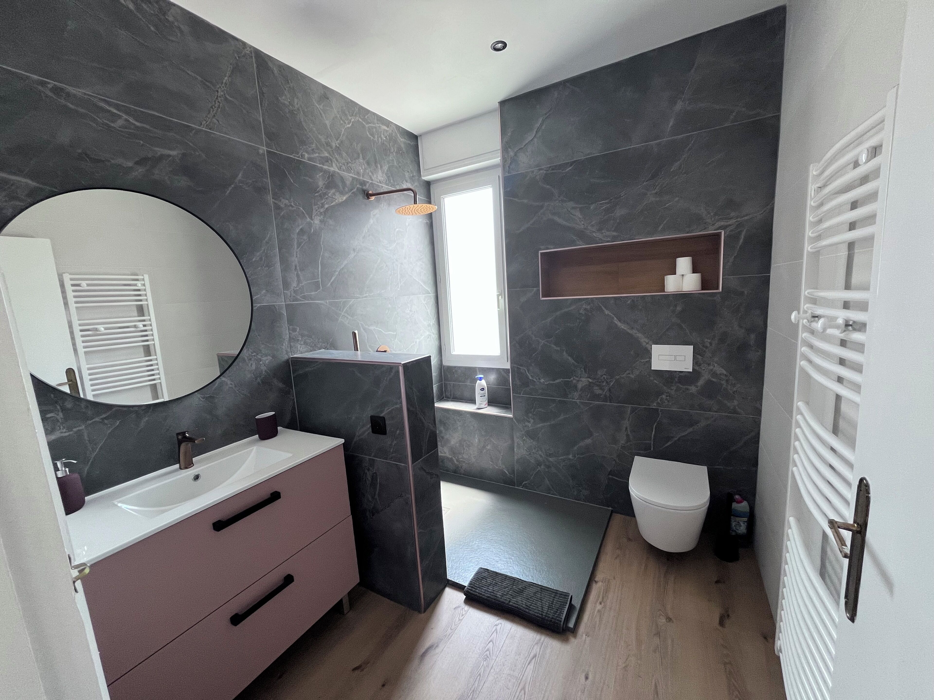 A modern bathroom with a marble wall, a sink with a mirror, a toilet, and a shower area.