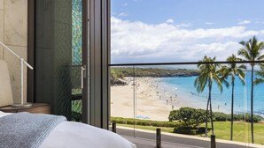 Habitación de hotel con un ventanal grande que da a una playa, palmeras y un cielo despejado.