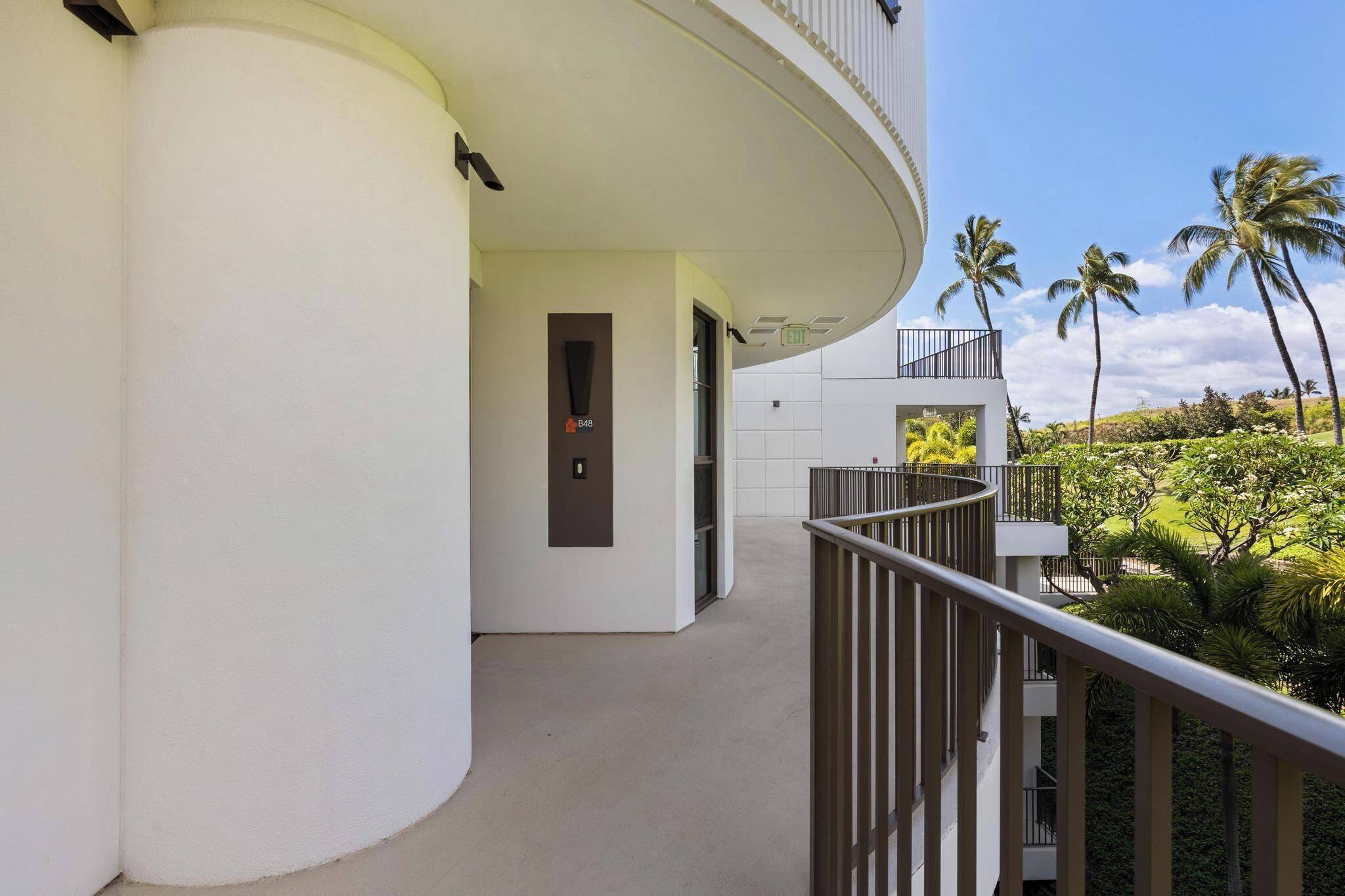 Un bâtiment moderne doté d’un balcon incurvé, d’une porte avec un numéro et offrant une vue sur des palmiers et une végétation luxuriante.