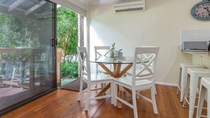 Un comedor con mesa de vidrio y sillas blancas, una puerta corredera de cristal que da a un jardín y un aire acondicionado de pared.