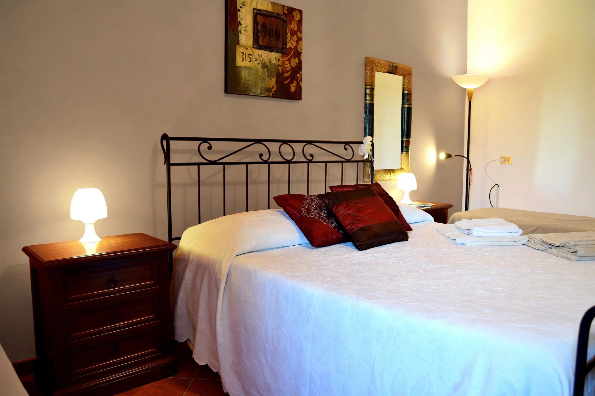 Cama bem arrumada com roupa de cama branca e vermelha, mesa de cabeceira de madeira com abajur, espelho fixado na parede e quadro acima da cama.
