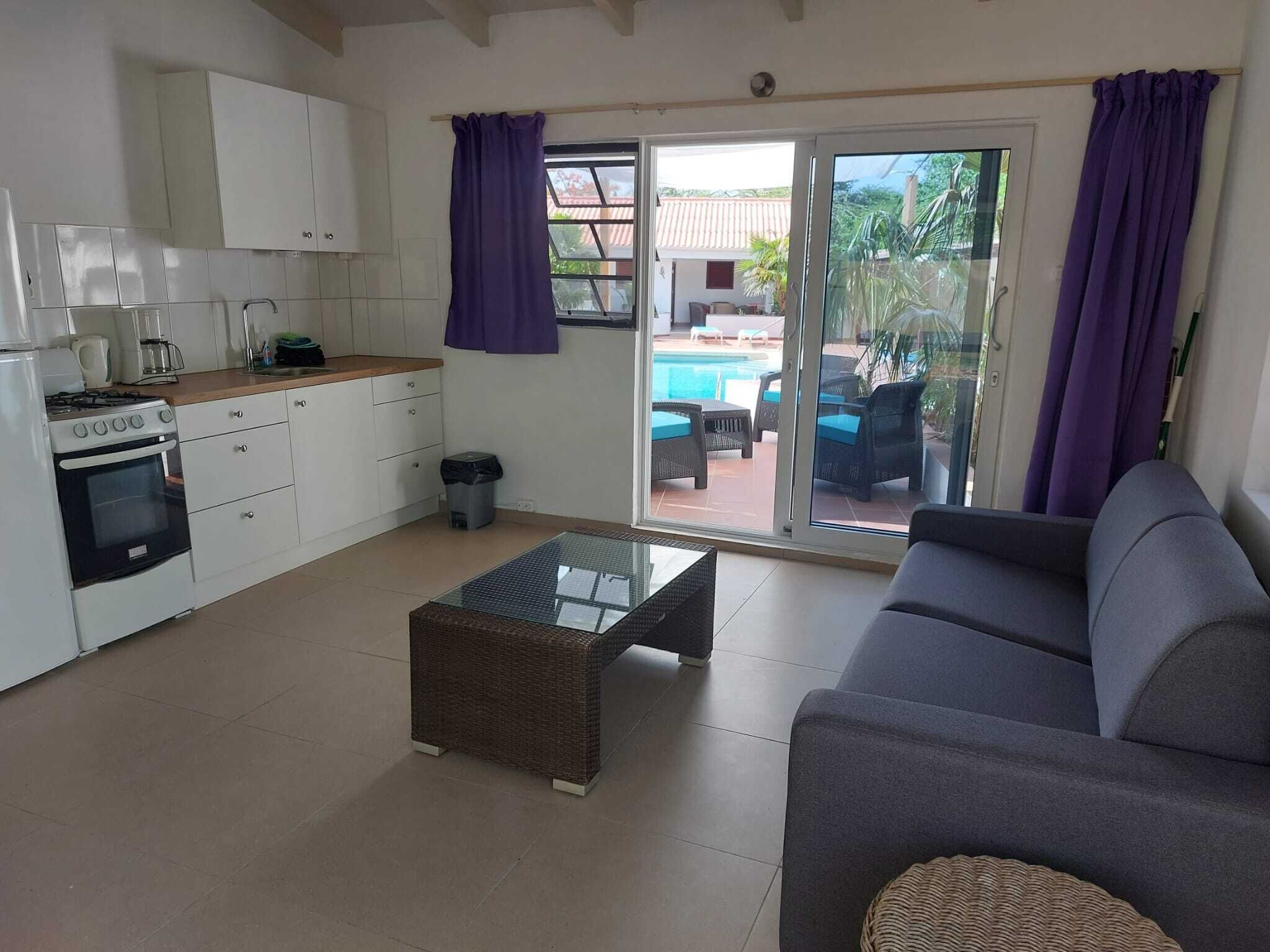 Een compact leefruimte met een kitchenette, een glazen salontafel, een bank en een glazen schuifdeur naar een buitengebied met een zwembad.
