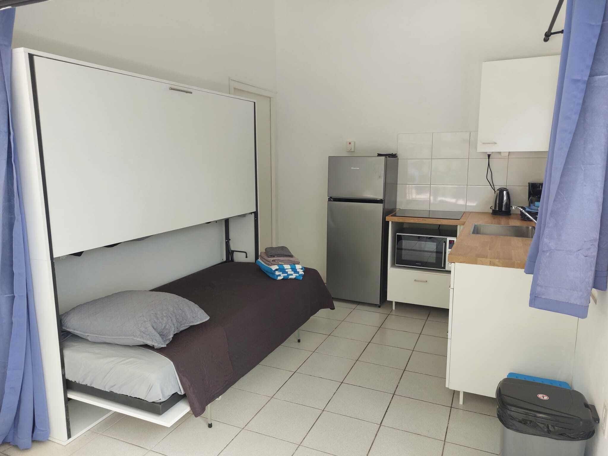 A compact room with a bed, a small kitchenette, and a trash bin.