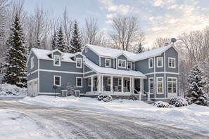 A two-story house with a porch and a garage, surrounded by snow-covered trees.