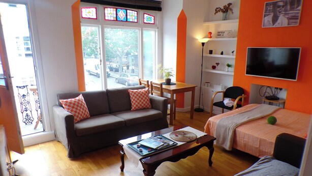 Un salon comprenant un canapé, une table basse, un téléviseur et un lit. Il y a une fenêtre donnant sur la rue.