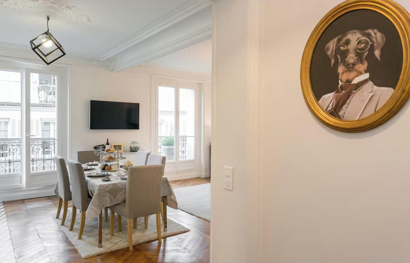 Un comedor con una mesa dispuesta para cuatro, un televisor montado en la pared y un cuadro enmarcado de un perro en la pared.