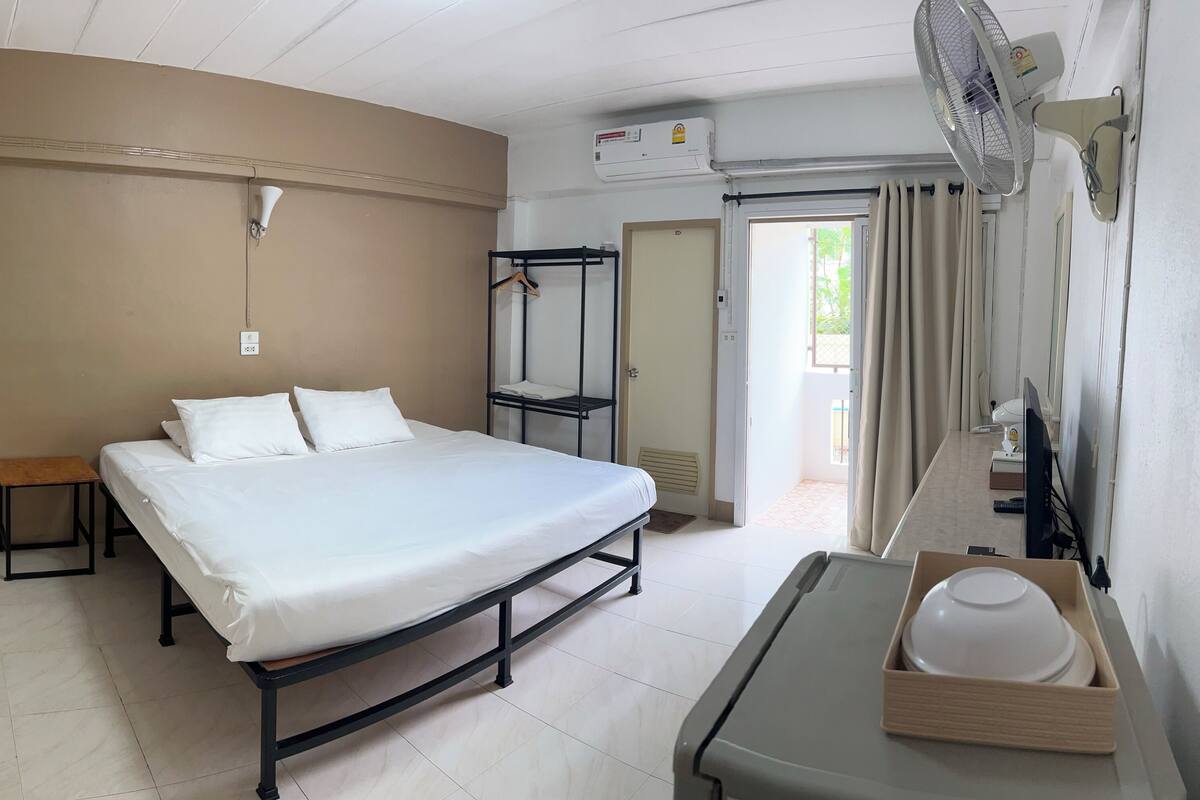 Habitación de hotel con cama, mesita de noche y escritorio con computadora. Hay una ventana con cortinas, unidad de aire acondicionado y una puerta que conduce a otra habitación.