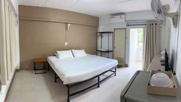 A hotel room with a bed, bedside table, and a desk with a computer. There is a window with curtains, an air conditioning unit, and a door leading to another room.