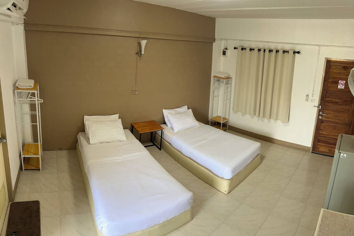 Una habitación de hotel con dos camas individuales, una mesita, un estante con un ventilador y una puerta con un cartel.