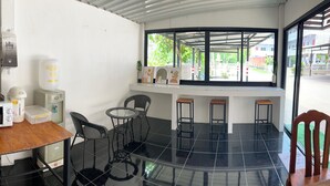 A compact kitchen with a microwave, toaster, and coffee maker on a wooden table. There is a small round table with chairs and a window with a view of the outside. - Phuviewplace (Chiang Kham)