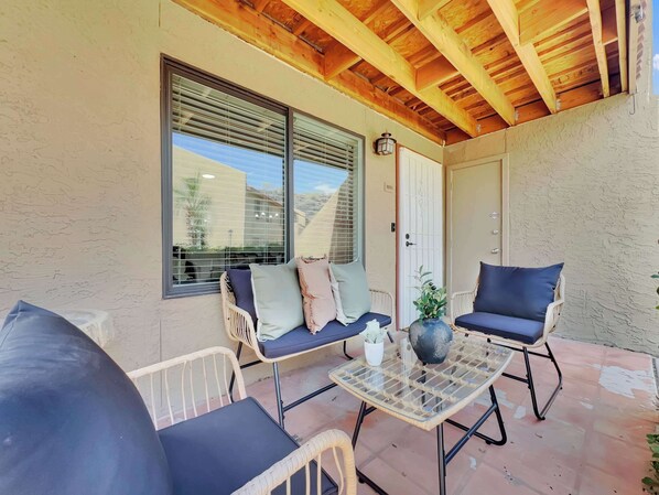 Une terrasse extérieure aménagée avec des meubles en osier, une table en verre et une vue sur un bâtiment, visible à travers de grandes fenêtres.