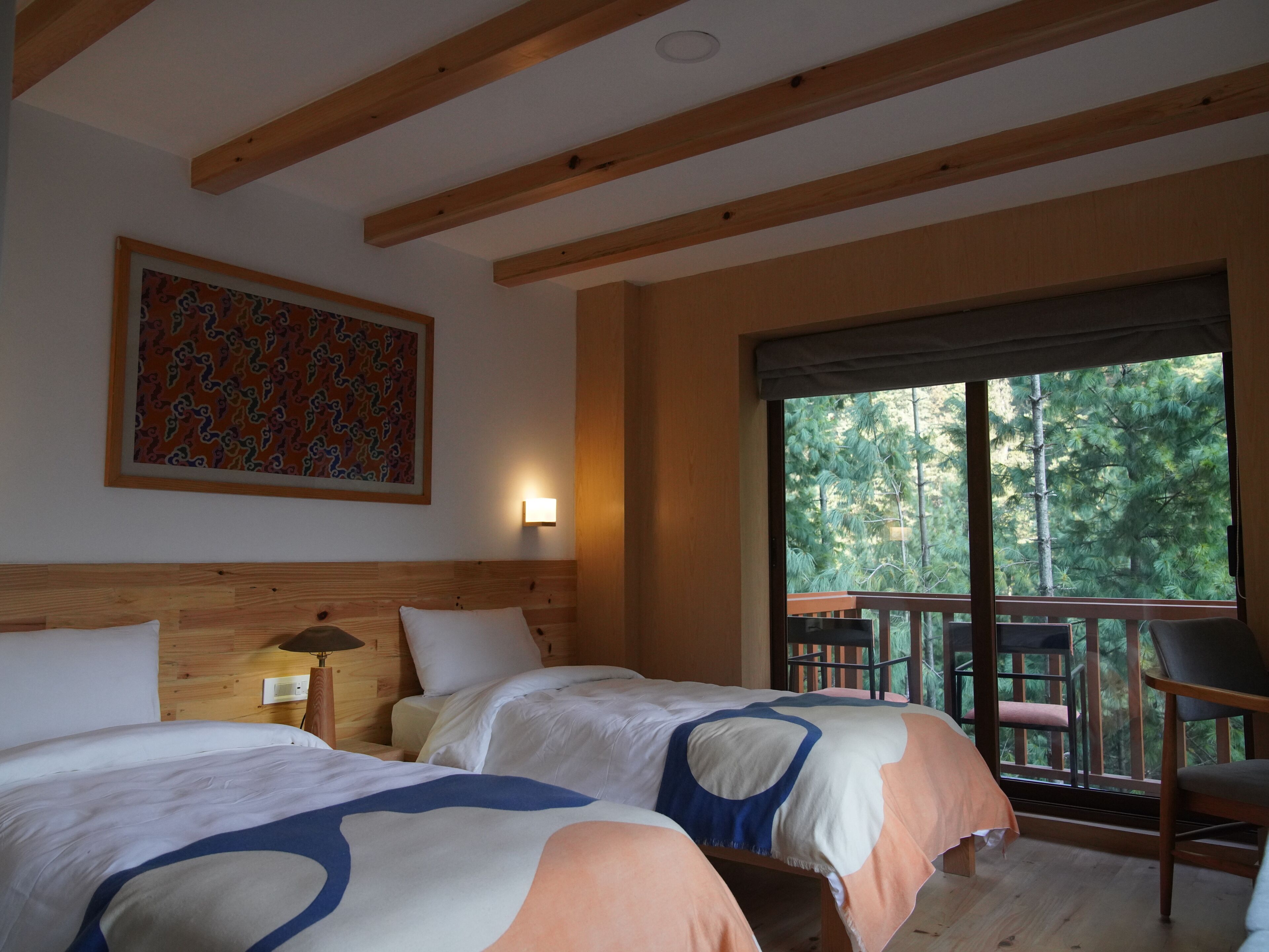 Quarto com duas camas, teto de madeira, um quadro na parede, janela com vista para árvores e uma varanda com cadeira.