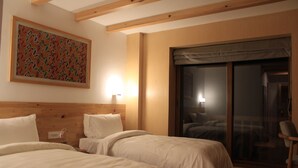 Ein Hotelzimmer mit zwei Betten, einem Holzkopfstück, einem Gemälde an der Wand, einer Schiebetür aus Glas und einem Sessel.